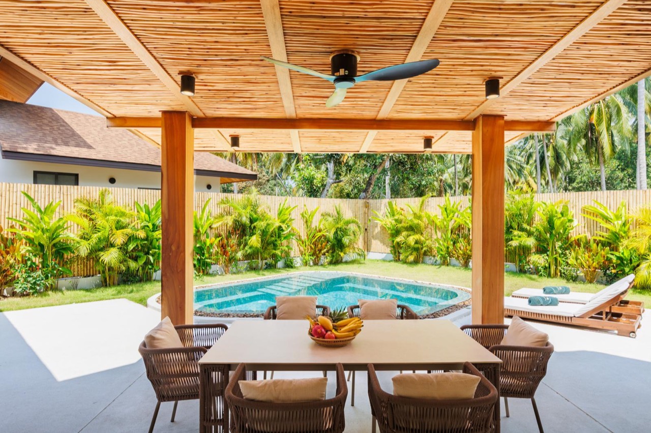 Long dining table in the shade beside the pool, with a bamboo roof and tropical garden